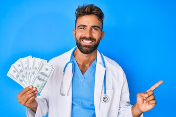 Young hispanic man wearing doctor uniform holding dollars smiling happy pointing with hand and finger to the side