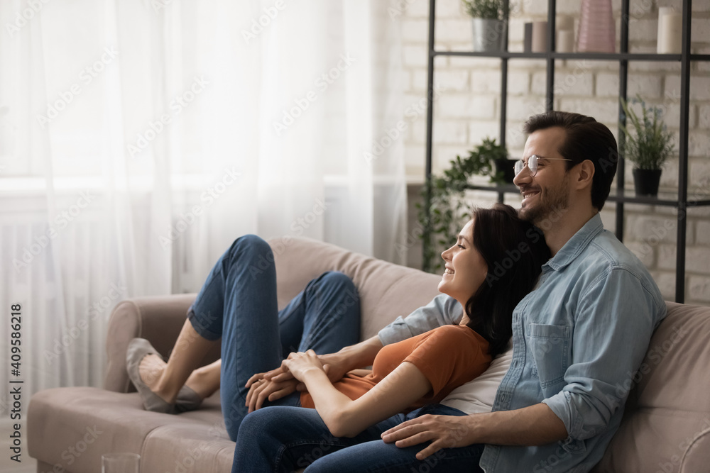 Happy dreamy young couple hugging, relaxing on cozy couch together, renters enjoying leisure time in new apartment, smiling wife and husband planning future, sitting on comfortable sofa at home - obrazy, fototapety, plakaty 