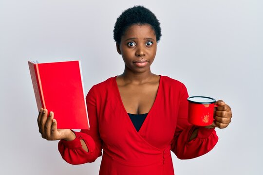 Young african woman with short hair reading a book and drinking a cup of coffee clueless and confused expression. doubt concept.