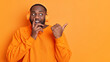 Positive black unshaven man with thick beard points thumb away on blank space has good mood listens audio track via headphones dressed in long sleeved jumper poses against vivid orange background