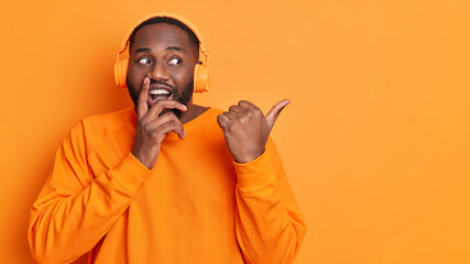 Positive black unshaven man with thick beard points thumb away on blank space has good mood listens audio track via headphones dressed in long sleeved jumper poses against vivid orange background