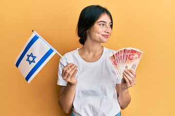 Wall Mural - Young caucasian woman holding israel flag and shekels banknotes smiling looking to the side and staring away thinking.