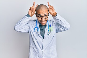 Wall Mural - Hispanic adult man wearing doctor uniform and stethoscope posing funny and crazy with fingers on head as bunny ears, smiling cheerful