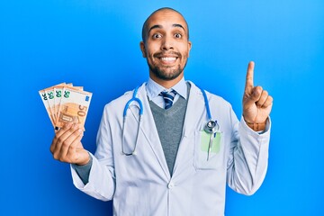 Sticker - Hispanic adult doctor man wearing medical uniform holding 50 euros banknotes smiling with an idea or question pointing finger with happy face, number one