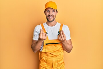 Canvas Print - Young handsome man wearing handyman uniform over yellow background doing money gesture with hands, asking for salary payment, millionaire business