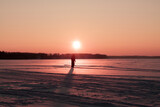 Fototapeta  - Horizontal landscape conceptual photo with a silhouette of alone human on the snow-covered field against setting sun over horizon during winter peachy sunset 