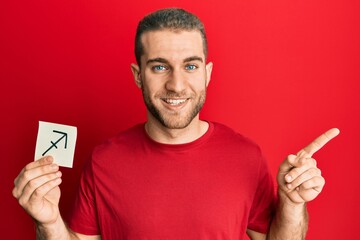 Poster - Young caucasian man holding paper with sagittarius zodiac sign smiling happy pointing with hand and finger to the side