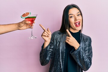 Wall Mural - Young girl wearing sexy party jacket and hispanic arm offering cocktail smiling and looking at the camera pointing with two hands and fingers to the side.