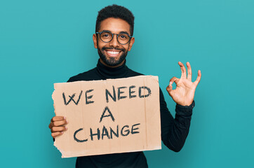 Wall Mural - Young african american man holding we need a change banner doing ok sign with fingers, smiling friendly gesturing excellent symbol