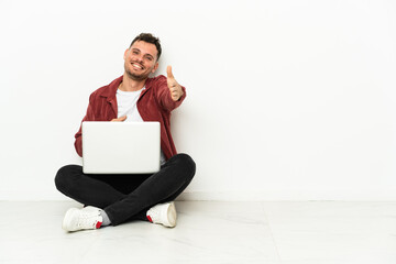 Wall Mural - Young handsome caucasian man sit-in on the floor with laptop shaking hands for closing a good deal