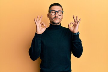 Wall Mural - Handsome man with tattoos wearing turtleneck sweater and glasses relaxed and smiling with eyes closed doing meditation gesture with fingers. yoga concept.