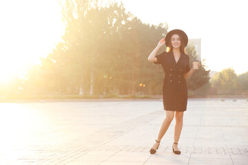 Canvas Print - Beautiful young woman in stylish black dress and hat with cup of coffee on city street