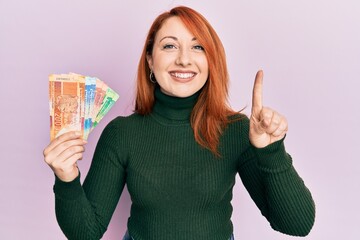 Sticker - Beautiful redhead woman holding south african rand banknotes smiling with an idea or question pointing finger with happy face, number one