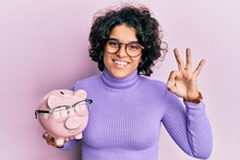 Young Hispanic Woman With Curly Hair Holding Piggy Bank With Glasses Doing Ok Sign With Fingers, Smiling Friendly Gesturing Excellent Symbol
