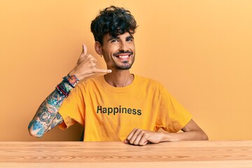 Wall Mural - Young hispanic man wearing tshirt with happiness word message sitting on the table smiling doing phone gesture with hand and fingers like talking on the telephone. communicating concepts.