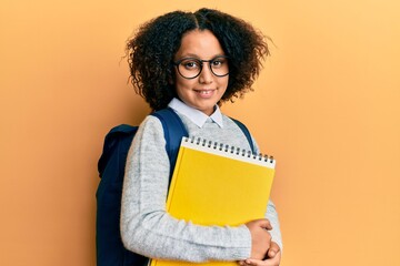 Sticker - Young little girl with afro hair wearing school bag and holding books looking positive and happy standing and smiling with a confident smile showing teeth