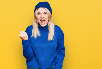 Wall Mural - Young caucasian woman wearing wool winter sweater and cap angry and mad raising fist frustrated and furious while shouting with anger. rage and aggressive concept.