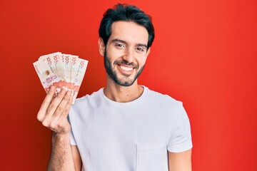 Sticker - Young hispanic man holding 10 colombian pesos banknotes looking positive and happy standing and smiling with a confident smile showing teeth