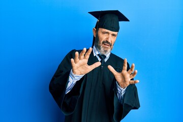 Poster - Middle age hispanic man wearing graduation cap and ceremony robe afraid and terrified with fear expression stop gesture with hands, shouting in shock. panic concept.