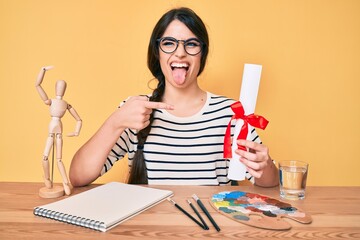 Wall Mural - Brunette teenager girl artist holding painter diploma sticking tongue out happy with funny expression.