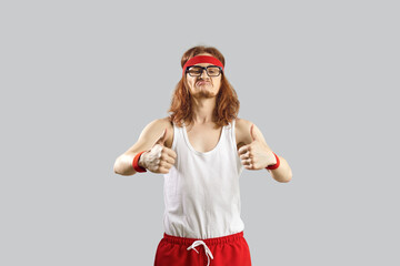 Funny nerdy guy giving you motivation to exercise. Skinny man in gym headband and tank top standing isolated on grey background and showing thumbs up gesture. Sports workout and fitness humour concept