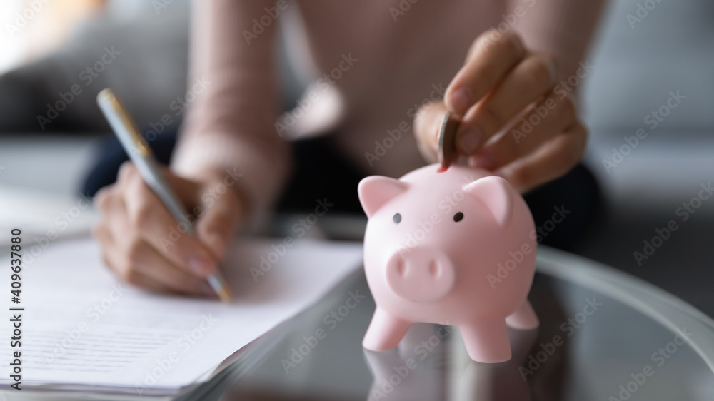 Close up young woman putting coin in piggy bank, writing household expenses on paper, planning domestic monthly bills payments or medical insurance services, managing savings or calculating taxes. - obrazy, fototapety, plakaty 