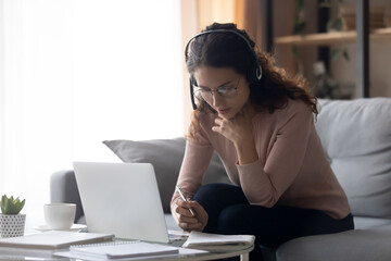 Wall Mural - Focused millennial woman in eyeglasses wearing headset with microphone, involved in studying on online lesson using computer, listening educational lecture, writing notes, e-learning concept.