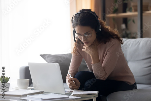 Focused millennial woman in eyeglasses wearing headset with microphone, involved in studying on online lesson using computer, listening educational lecture, writing notes, e-learning concept. © fizkes