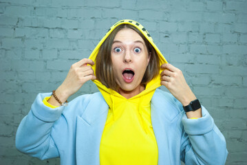 Closeup portrait of shocked girl with brown eyes and open mouse look at the camera holding yellow hood by both hand. Concept of advertising isolated on blue background