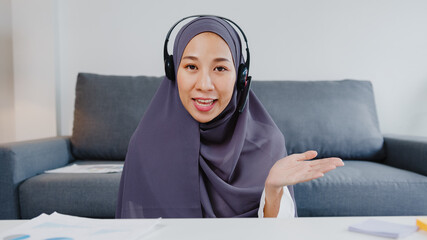 Canvas Print - Asia muslim lady wear headphone using computer laptop talk to colleagues about plan in video call meeting while remotely work from home at living room. Social distancing, quarantine for corona virus.