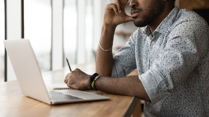 Wall Mural - Pensive young black man remote worker freelancer sit at work desk read study data on laptop screen. Thoughtful african guy student ponder on learning material hold pen prepare to make notes. Close up