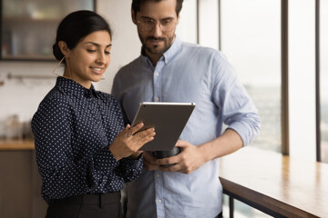 Canvas Print - Young hindu female office employee mentor show interested male intern problem solution online using tablet. Two coworkers of different ethnicities discuss work meeting at dining area on lunch break.