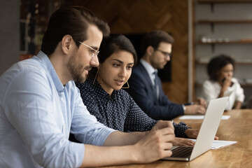 Canvas Print - Focused millennial male intern look at laptop screen listen to skilled indian female mentor. Hindu woman experienced worker consult young man new employee help in computer work at corporate network