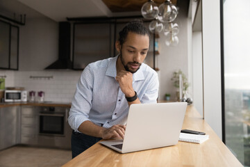 Thoughtful young african male stand by desk with laptop at home kitchen read important message email think on answer create respond. Focused biracial man student contact friend using messenger pc app
