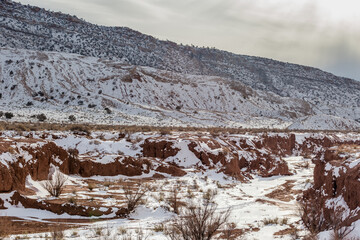 Red rock valley with barren bushes and mountainside covered in snow on overcast day in rural New Mexico