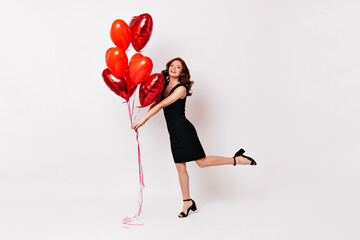 Wall Mural - Full length shot of ginger woman in black dress. Studio photo of slim girl dancing with heart shaped balloons.