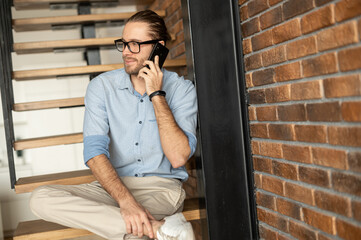 Handsome young hipster man wearing glasses and casual attire is busy, talking on mobile phone and smiling while sitting on the stairs in the house with the loft modern interior or in the coffee shop