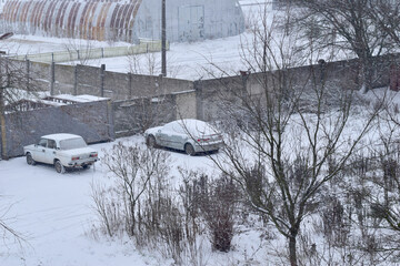 Two cars covered with snow stand near the fence.