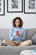 © insta_photos - Smiling latin teenage girl holding smartphone using mobile apps technology at home. Happy hispanic young woman texting, checking online social media applications sitting on couch in apartment.