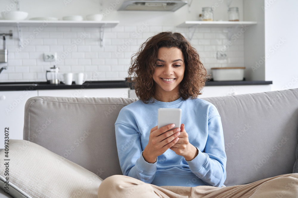 Happy hispanic teen girl looking at smartphone relaxing on couch at home, enjoying using online mobile apps technology, playing games on cell phone, checking messages or social media posts. - obrazy, fototapety, plakaty 