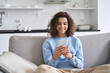 Happy hispanic teen girl looking at smartphone relaxing on couch at home, enjoying using online mobile apps technology, playing games on cell phone, checking messages or social media posts.