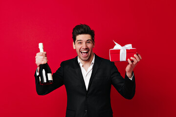 Happy man with wine and present box smiling at camera. Front view of brunette boy with gift isolated on red.