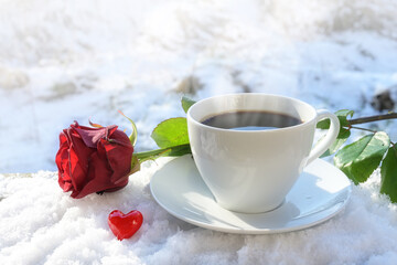 Hot coffee in a white cup served outdoors in the snow, decorated with a red rose and a little heart from glass on a sunny winter day, love concept for a happy valentines day, copy space