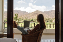 Freelancer, Remote Work, Work From Home. Young Woman Is Working On A Laptop On Her Balcony Overlooking The Tropical Garden