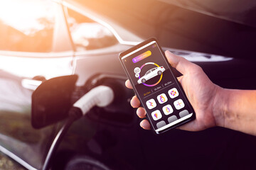 Using mobile smartphone device refueling electric vehicle EV car mock UI application display screen, hand holding cellphone at electric car charging station connected power cable alternative energy