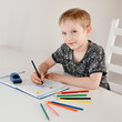 © Елена Лавринович - a 7-year-old boy of European appearance draws in a notebook with pencils. The child at the table looks at the camera. Drawing training, creativity development