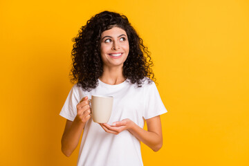 Wall Mural - Photo portrait of curly brunette keeping cup with hot beverage looking empty space smiling isolated on vibrant yellow color background