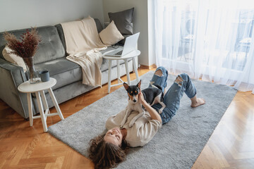 Wall Mural - Pretty curly happy young woman smiling while lying at home on carpet with her dog