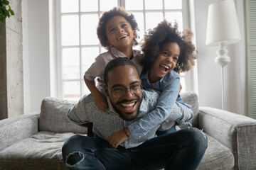 Wall Mural - Close up overjoyed African American man wearing glasses piggy backing kids, sitting on couch at home, excited laughing dad carrying adorable little son and daughter, family having fun, funny activity