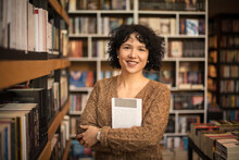 Portrait Of Smiling In Library Holding A Book. Looking At Camera.